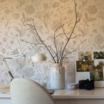 A cozy home office space is graced with Toile De Jouy Floral Wallpaper, where delicate beige florals provide a classic and refined backdrop. A desk with a white lamp, a vase with branches, and a collage of floral images complete the serene workspace.