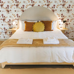 A bedroom elegantly decorated with Autumn Floral Wallpaper, featuring large floral prints in shades of rose and beige. The cozy setting includes a bed with white linens, mustard yellow pillows, and a round yellow cushion, creating a warm, inviting atmosphere.