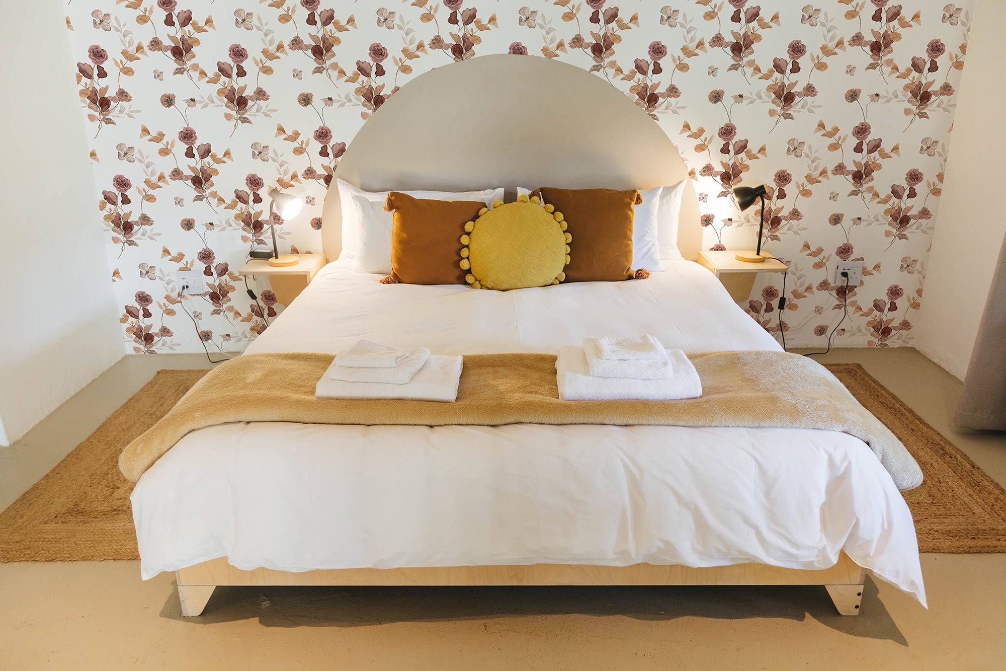 A bedroom elegantly decorated with Autumn Floral Wallpaper, featuring large floral prints in shades of rose and beige. The cozy setting includes a bed with white linens, mustard yellow pillows, and a round yellow cushion, creating a warm, inviting atmosphere.
