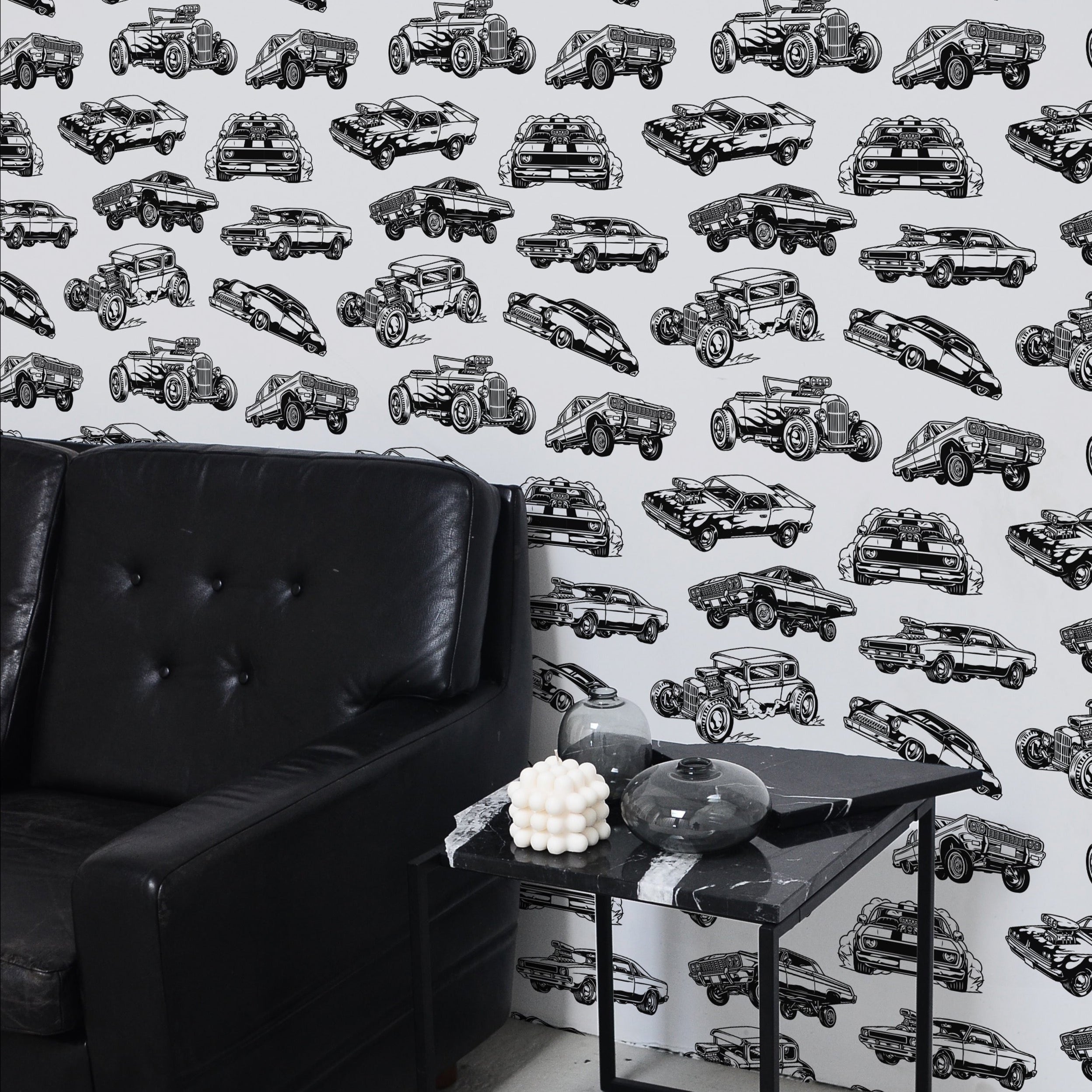 A contemporary living room setup with Vintage Cars Wallpaper as the focal wall. The decor includes a black leather sofa and a sleek black coffee table, complementing the monochrome car prints and creating a modern yet retro vibe.
