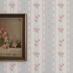 A section of wall adorned with Vintage Cutie Wallpaper featuring a pattern of pink roses aligned in vertical stripes against a pale beige background with delicate lace and line patterns. A classic framed painting of pink flowers complements the vintage aesthetic.