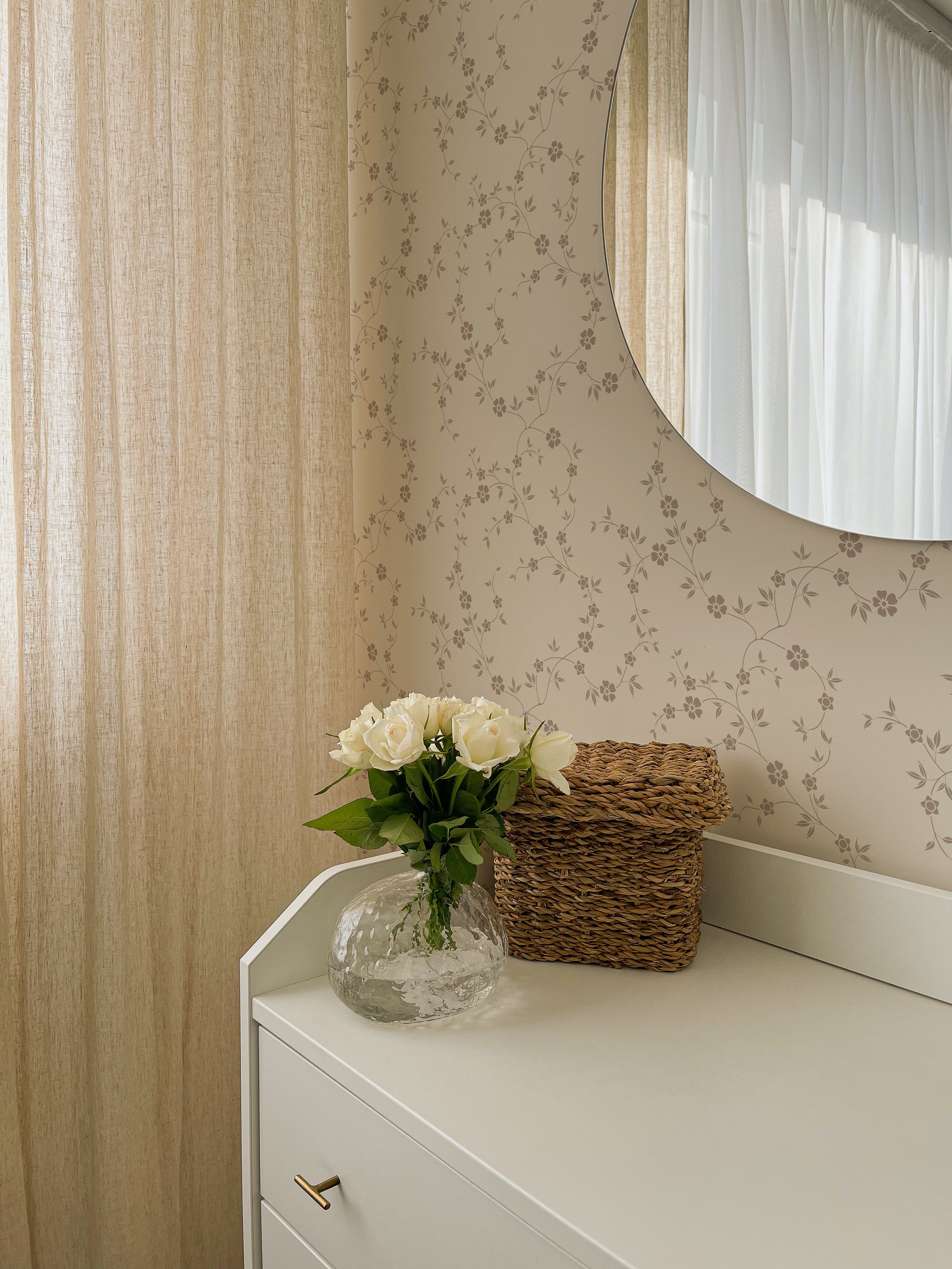 A chic corner of a room with a round mirror reflecting 'Charming Floral Wallpaper' on the adjacent wall. The wallpaper's beige floral pattern adds a warm, inviting feel to the space, complemented by a vase of white roses and a woven basket.