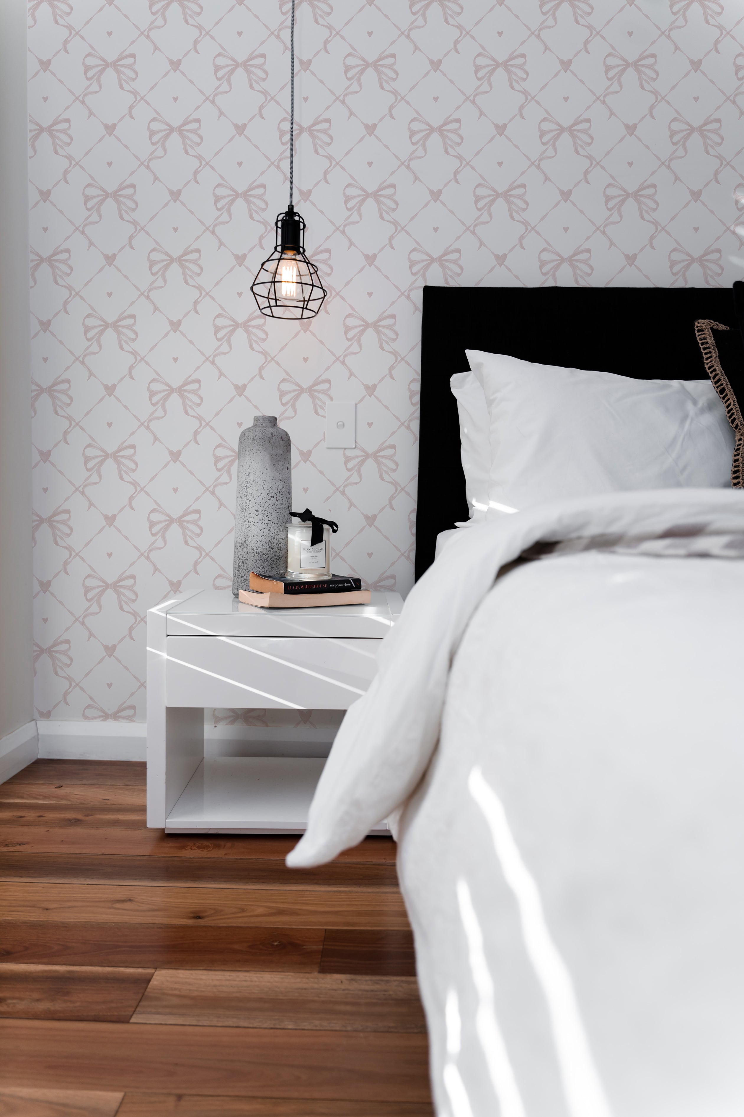 A modern bedroom featuring Delicate Watercolour Bows Wallpaper with a pattern of light pink bows and hearts on a white background. The elegant wallpaper enhances the room's aesthetic, complemented by a black headboard, white bedding, and a minimalist bedside table.