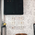 A cozy corner of a room featuring a wooden chair in front of a wall adorned with a floral-patterned wallpaper. The wallpaper showcases a delicate, hand-drawn design of wildflowers in black sketch-like strokes on an off-white background. Above the chair hangs a series of small framed illustrations of coffee cups with whimsical faces. To the left, a tall black pedestal holds a vase with yellow flowers, enhancing the room's botanical theme.