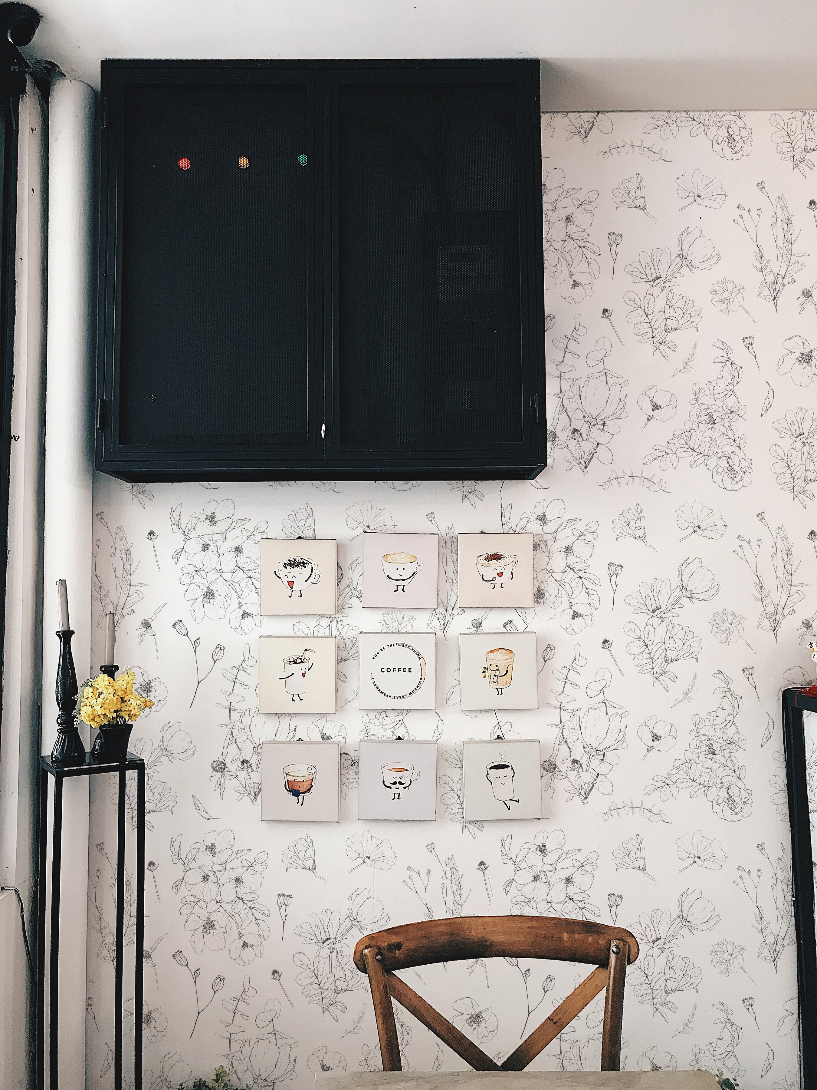 A cozy corner of a room featuring a wooden chair in front of a wall adorned with a floral-patterned wallpaper. The wallpaper showcases a delicate, hand-drawn design of wildflowers in black sketch-like strokes on an off-white background. Above the chair hangs a series of small framed illustrations of coffee cups with whimsical faces. To the left, a tall black pedestal holds a vase with yellow flowers, enhancing the room's botanical theme.
