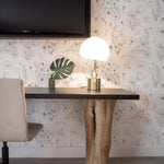 A modern workspace setup against a backdrop of Wildflower Sketch Wallpaper, which displays a minimalistic black-on-white floral design. The workspace includes a sleek, dark wooden desk supported by a unique, organic-shaped, cream-colored base that resembles a tree trunk. A contemporary round lamp with a frosted glass shade sits on the desk, accompanied by a monstera leaf in a green glass vase, and the room is completed with a flat-screen TV mounted on the wall above.