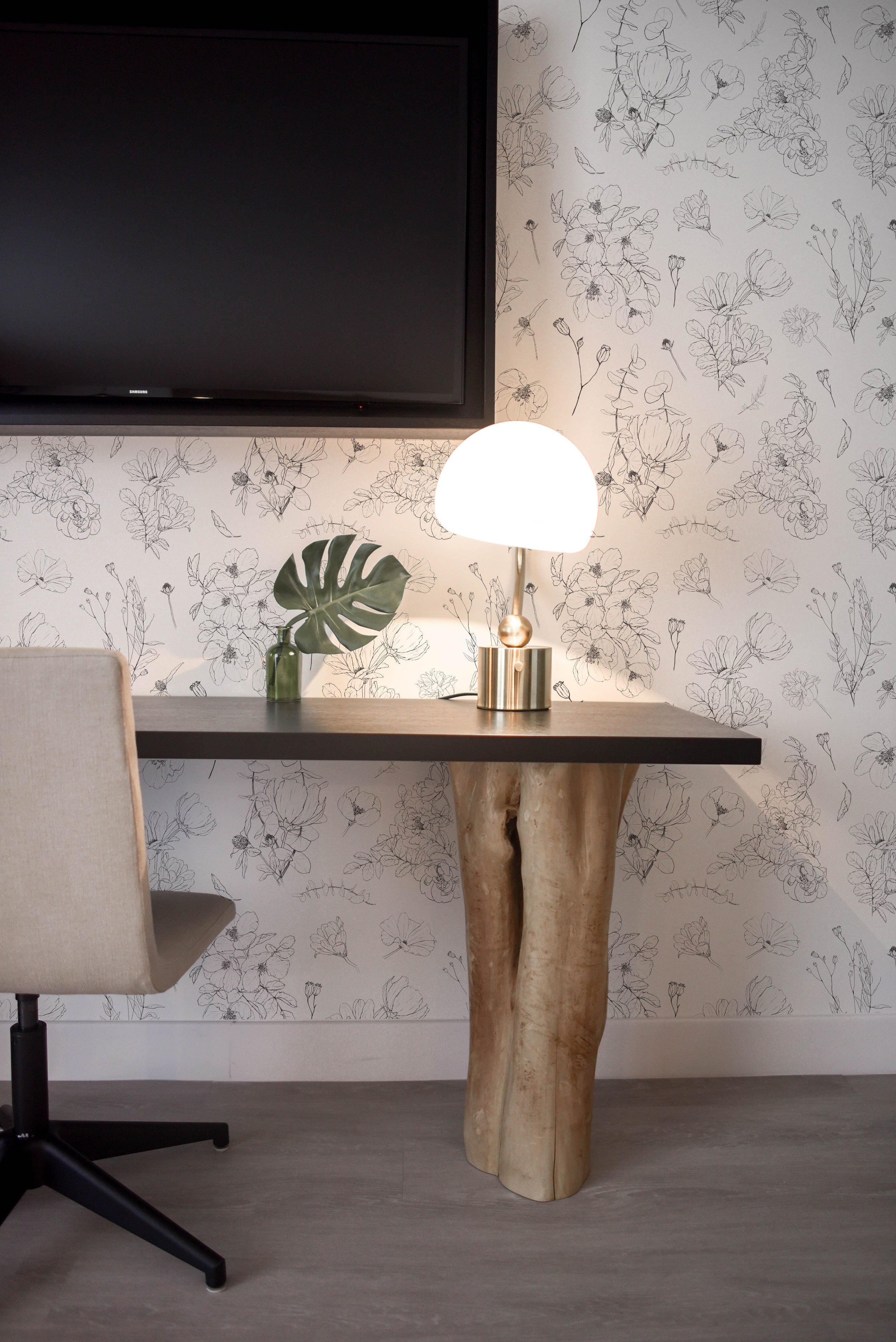 A modern workspace setup against a backdrop of Wildflower Sketch Wallpaper, which displays a minimalistic black-on-white floral design. The workspace includes a sleek, dark wooden desk supported by a unique, organic-shaped, cream-colored base that resembles a tree trunk. A contemporary round lamp with a frosted glass shade sits on the desk, accompanied by a monstera leaf in a green glass vase, and the room is completed with a flat-screen TV mounted on the wall above.