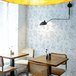 An inviting cafe scene with tables against a wall covered in Wildflower Sketch Wallpaper. The black and white floral sketches provide a charming backdrop for the modern, yet cozy dining space. A striking yellow pendant light casts a warm glow over the wooden tables and cane-backed chairs, creating an ambiance that is both stylish and relaxed.