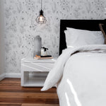 An elegant bedroom corner showcasing a black upholstered headboard against a wall covered in 'Wildflower Sketch Wallpaper,' with the intricate black-and-white floral designs complementing the simple white nightstand and industrial-style hanging light fixture.