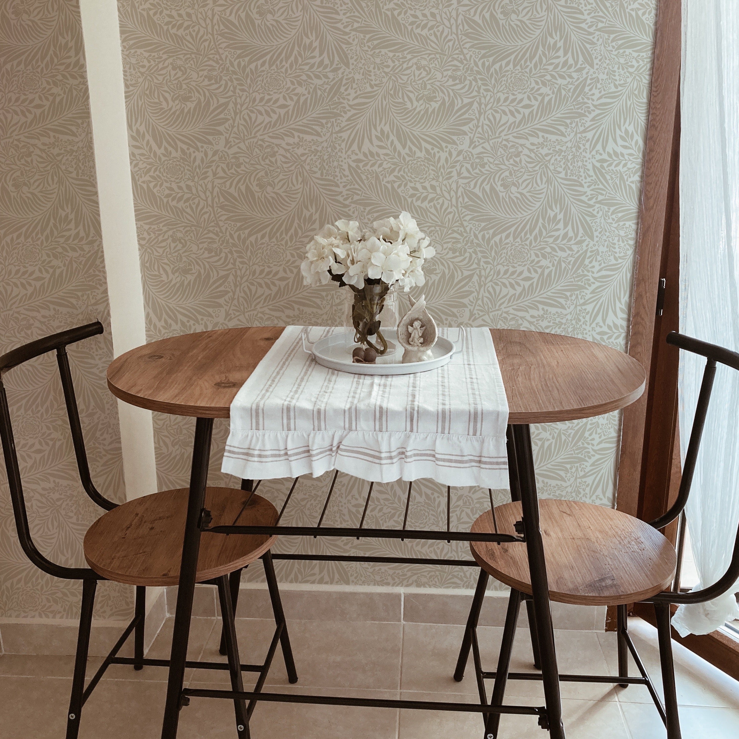  quaint dining area graced by the 'William Bough Wallpaper', featuring a round wooden table with black chairs, set under a simple white cloth and a vase of white flowers