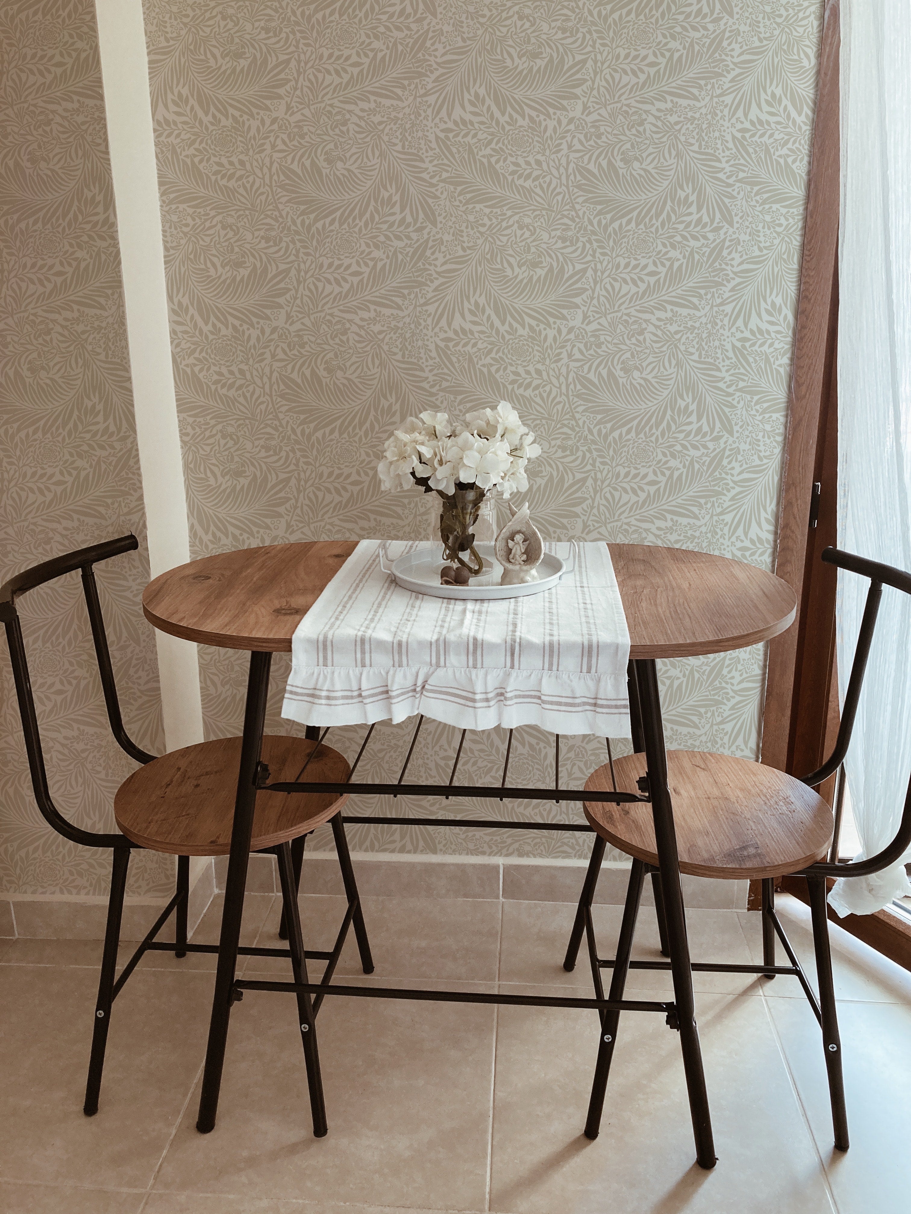 quaint dining area graced by the 'William Bough Wallpaper', featuring a round wooden table with black chairs, set under a simple white cloth and a vase of white flowers