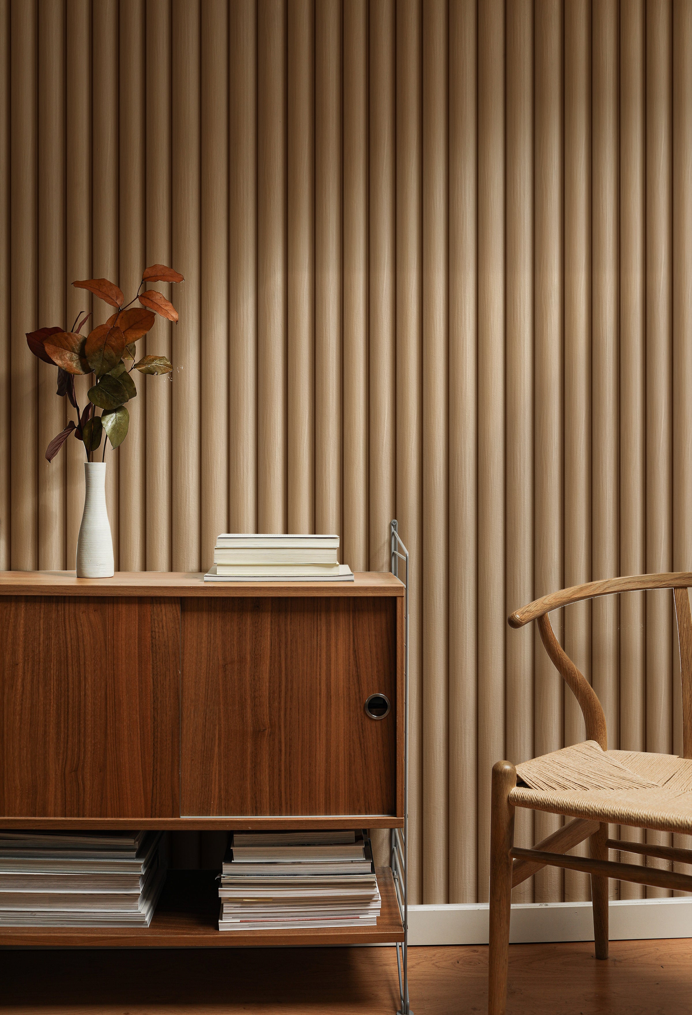 A stylish room with Wooden Pillar Wallpaper showcasing vertical grooves in a warm wood tone. The wallpaper enhances the room's aesthetic, highlighted by a mid-century modern wooden cabinet with books and a vase with autumn leaves.