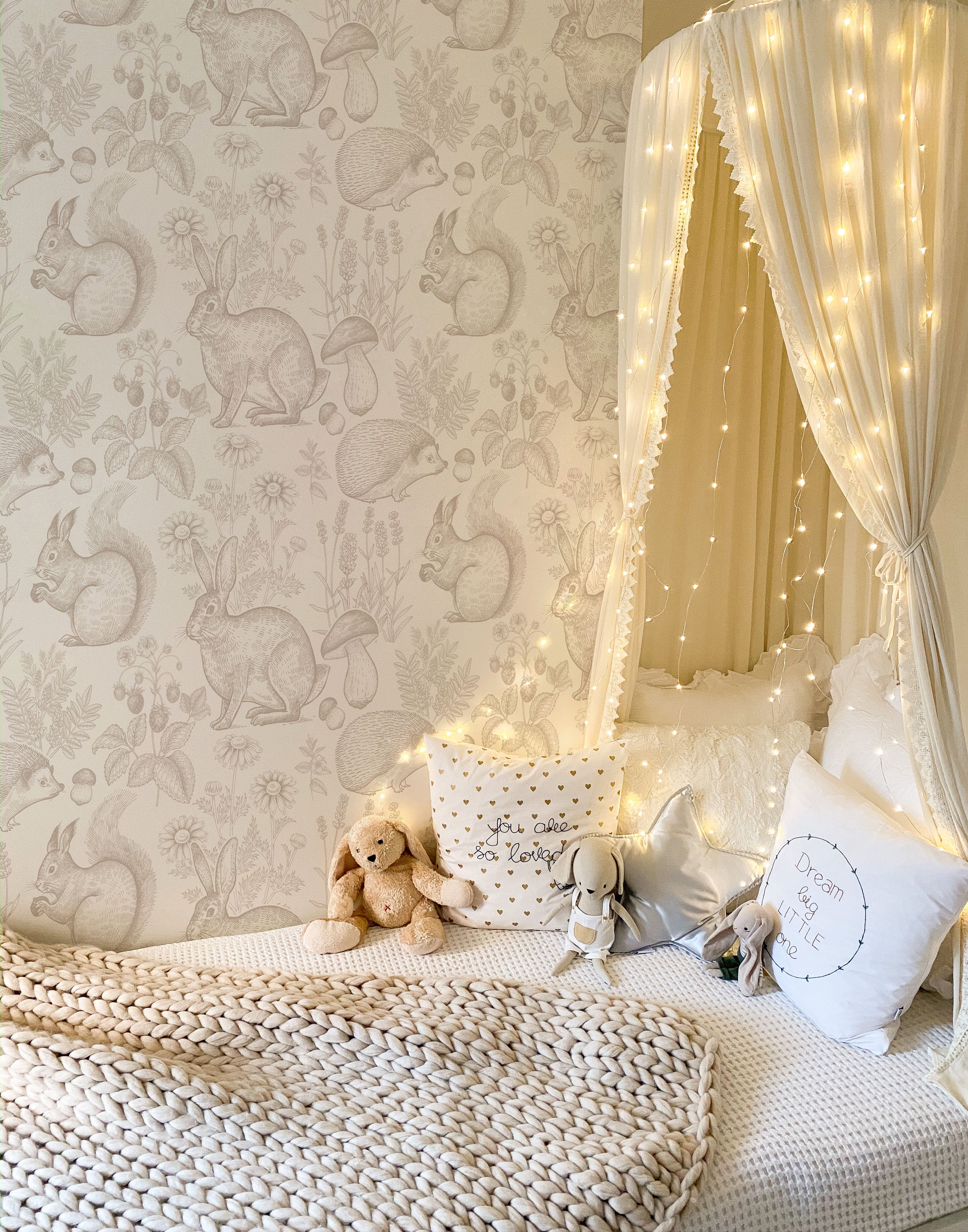 A cozy children’s nook adorned with the "Woodland Creatures Wallpaper - Beige," featuring a charming array of hand-drawn forest animals like rabbits, hedgehogs, and squirrels. The scene is complete with a bed draped in fairy lights, fluffy pillows, and plush toys, creating a whimsical and inviting space for a child.