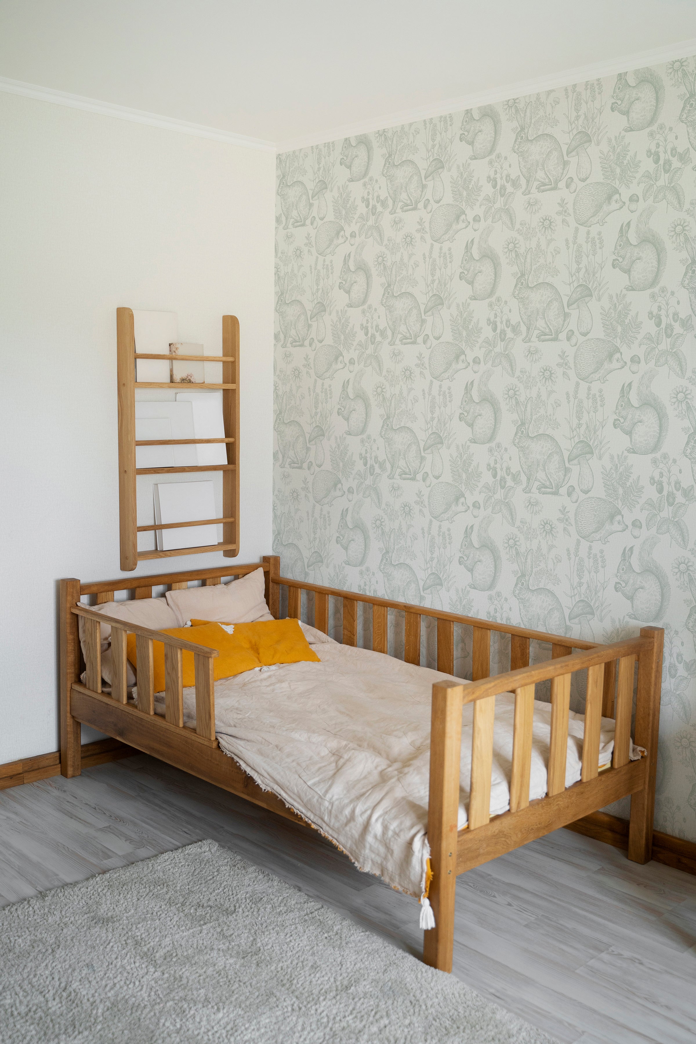 A child's bedroom features the Woodland Creatures Wallpaper - Light Sage, with illustrations of rabbits, hedgehogs, squirrels, and floral elements creating a peaceful, nature-inspired wall. A wooden bed and ladder shelf complement the gentle sage color of the wallpaper.