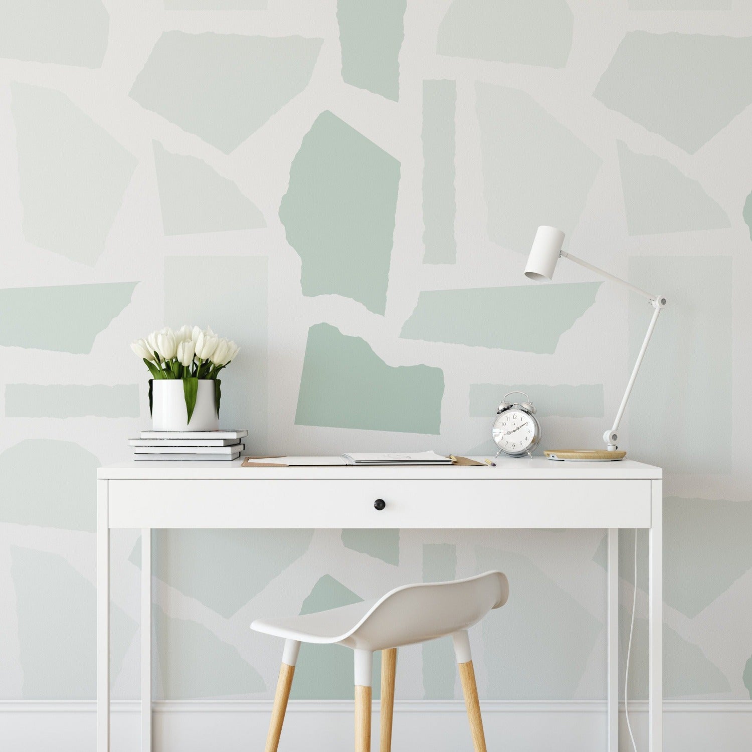 A photo of a modern office setup with a white desk and chair against a wall covered in the abstract green and white collage wallpaper. The desk is accessorized with a white table lamp, a small clock, a vase of white flowers, and a stack of books.