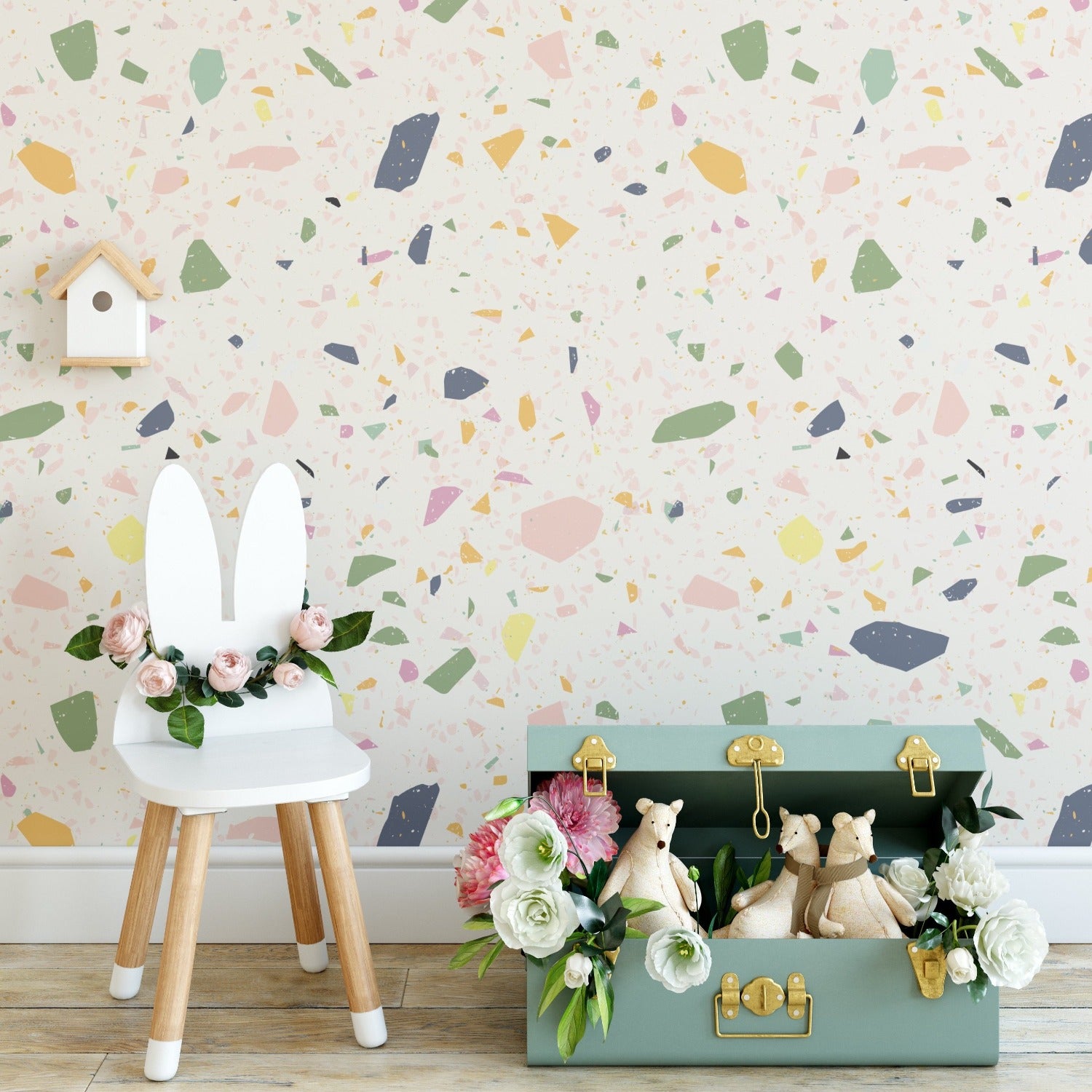 A styled children's room corner with the Cotton Candy Terrazzo wallpaper in the background, adorned with pastel confetti-like patterns. In the foreground, there's a chair with rabbit ears and a storage suitcase with stuffed animal toys and floral decorations, contributing to a playful and inviting atmosphere.