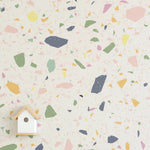 A close-up of a terrazzo-style wallpaper with a playful, pastel color scheme featuring large flecks of yellow, pink, green, and navy on a light cream background. A small, simplistic wooden birdhouse with a round entrance is centered at the bottom of the image, adding a quaint touch to the wallpaper's design.