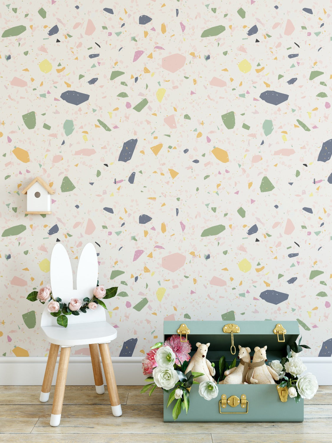 A styled children's room corner with the Cotton Candy Terrazzo wallpaper in the background, adorned with pastel confetti-like patterns. In the foreground, there's a chair with rabbit ears and a storage suitcase with stuffed animal toys and floral decorations, contributing to a playful and inviting atmosphere.