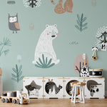 A playful and charming children's room with 'Forest Critters Kids Wallpaper' on the wall, featuring whimsical illustrations of woodland animals and plants in soft colors, above a child's storage unit filled with toys.