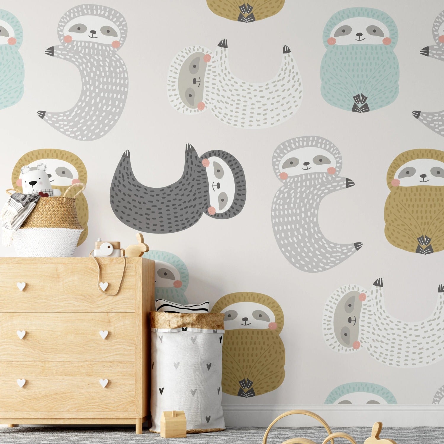 A wider view of the child's room showing more of the "Sleepy Sloths Wallpaper". The perspective includes a playful teepee-style tent in grey and black, with a cushion that has a similar sloth motif. The room is accessorized with wooden toys on the floor, a storage bin with a heart motif, and a few toys peeking out from the top of the dresser. The overall ambiance is cozy and conducive to a child's play and rest.