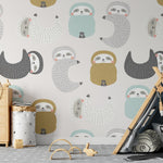 A wider view of the child's room showing more of the "Sleepy Sloths Wallpaper". The perspective includes a playful teepee-style tent in grey and black, with a cushion that has a similar sloth motif. The room is accessorized with wooden toys on the floor, a storage bin with a heart motif, and a few toys peeking out from the top of the dresser. The overall ambiance is cozy and conducive to a child's play and rest.