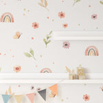 A child's room corner showing Rainbows and Flowers Wallpaper, with colorful bunting, decorative wooden blocks spelling HOME, and a white shelf against the floral and rainbow design.