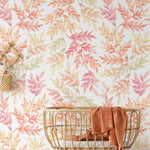A styled room scene featuring the "Bright Leaf Wallpaper" on the wall. The wallpaper has a pattern of watercolor leaves in soft shades of pink, yellow, and green, complementing the warm, natural decor elements in the room, including a gold wire basket and a terracotta-colored towel.