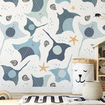 A playful underwater-themed wallpaper, Kids Wallpaper - Smiley Rays, featuring smiling stingrays and dotted fish in shades of blue and beige, interspersed with starfish and whimsical water swirls. The scene creates a happy aquatic atmosphere in a child's playroom with toys and a teepee shelf.
