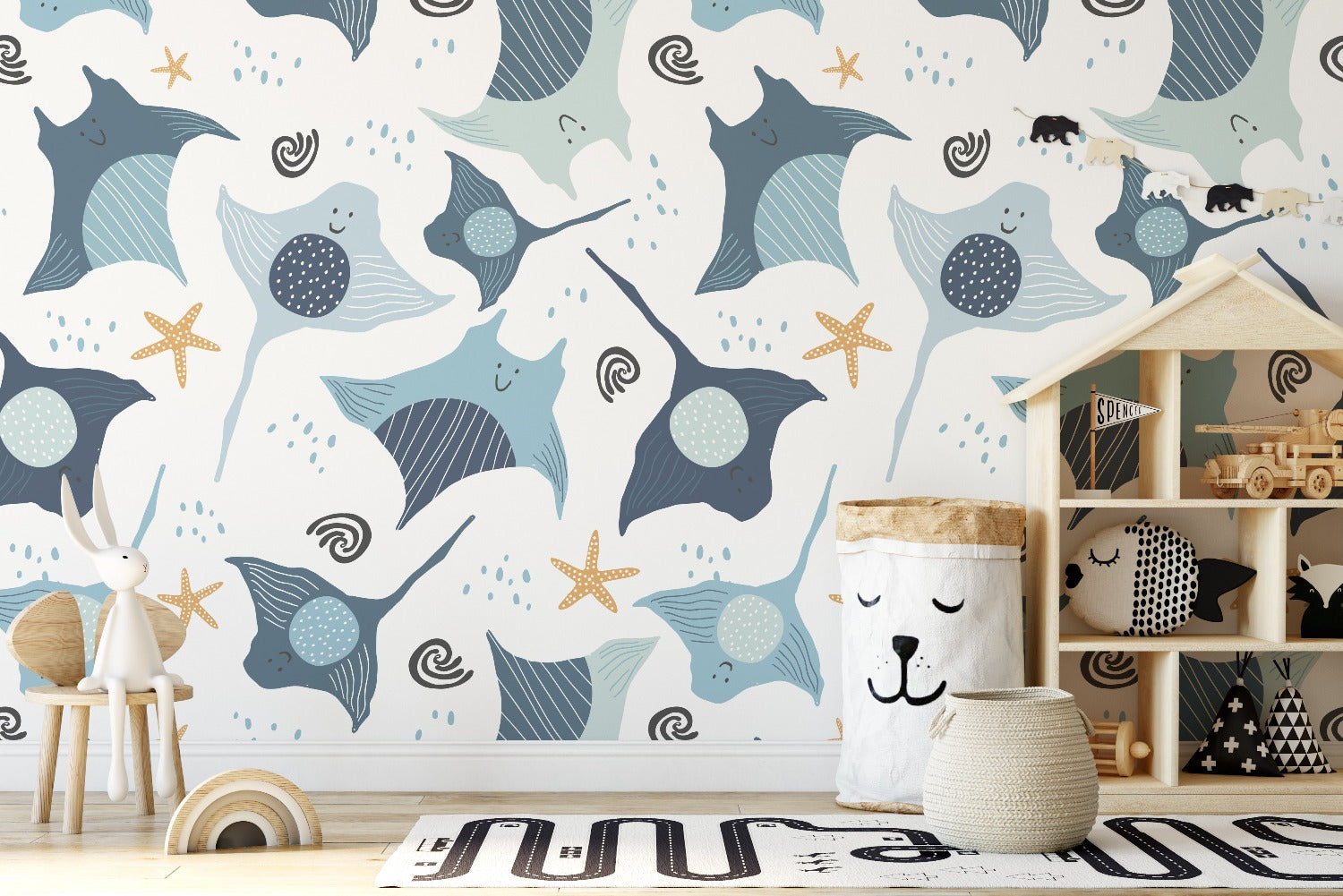 A playful underwater-themed wallpaper, Kids Wallpaper - Smiley Rays, featuring smiling stingrays and dotted fish in shades of blue and beige, interspersed with starfish and whimsical water swirls. The scene creates a happy aquatic atmosphere in a child's playroom with toys and a teepee shelf.