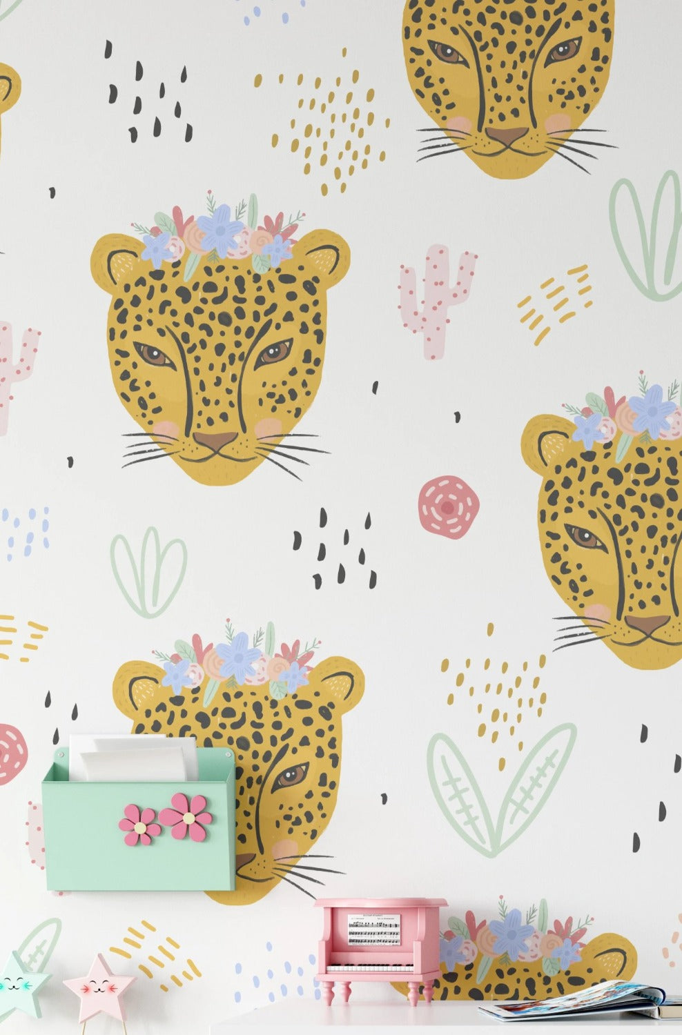 A room decorated with the same children's wallpaper featuring yellow leopard heads with flower crowns. The wallpaper covers the wall behind a white desk with pink and green accessories, creating a lively and cheerful atmosphere.