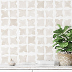 A styled interior space showcasing Moroccan Tile - Squares wallpaper with a subtle beige and white color scheme. The wallpaper features a repeating pattern of abstract square shapes resembling stylized butterflies or four-petal flowers. In the foreground, there's a potted fiddle-leaf fig tree next to a white console with a decorative shell and a stack of books, adding a touch of organic warmth to the room.