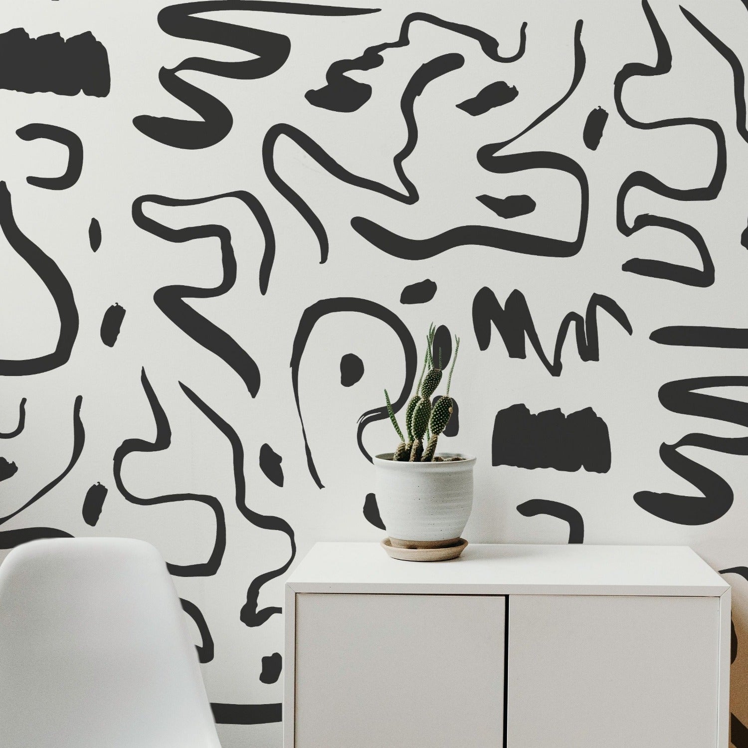 Modern and minimalist interior with a simple white cabinet and a cactus plant, set against a wall covered with Abstract Fusion Wallpaper. The wallpaper displays an intricate black on white abstract pattern, adding a bold artistic touch to the space.
