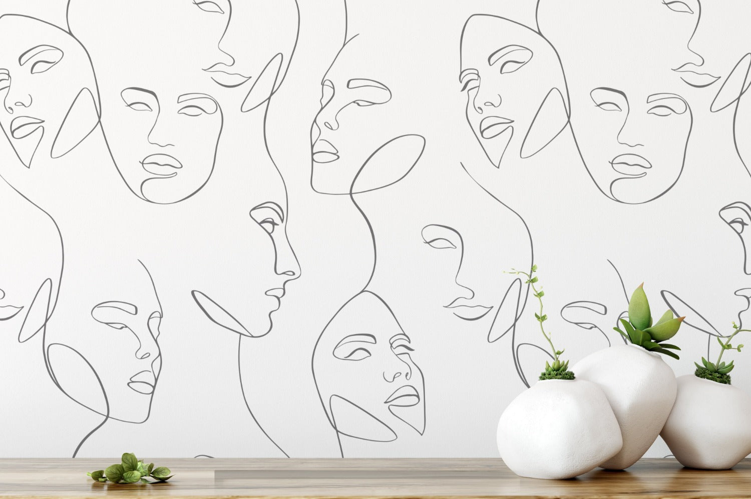 The Abstract and Minimal Wallpaper - Line Art III features a continuous line drawing of serene faces in black on a white background. A wooden surface with simplistic white vases holding green plants adds a natural touch to the modern aesthetic