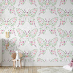 A room with a wall decorated with a playful wallpaper pattern featuring symmetrical butterfly shapes made of watercolor flowers and leaves in soft pinks, purples, and greens, creating a whimsical and airy atmosphere suitable for a child's nursery or playroom.
