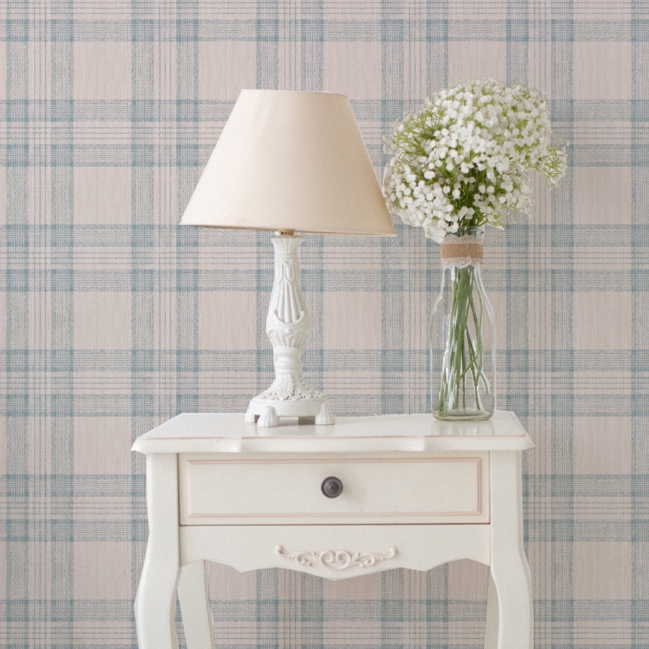 A quaint corner featuring the 'Plaid Wallpaper - Tartan Sand and Sky' as a backdrop, with a classic white side table bearing a traditional lamp with a beige shade and a clear glass vase filled with delicate white baby's breath flowers. The wallpaper's soft plaid pattern complements the vintage charm of the decor, creating a serene and welcoming space.