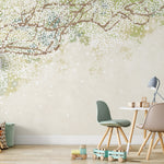 A charming children's playroom with a small table and chairs, soft toys, and a bookshelf against a wall decorated with the Sakura Serenade mural wallpaper. The mural showcases cherry blossom branches with white flowers on a soft green and beige background, creating a whimsical and soothing atmosphere for playtime.