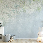 A whimsical nursery room corner featuring the Nursery Mural Wallpaper - Cherry Blossom Canopy, with a soothing illustration of cherry blossom branches in full bloom against a soft, muted blue background. A large, illuminated rabbit lamp sits beside a white cabinet decorated with playful plush toys and children's books