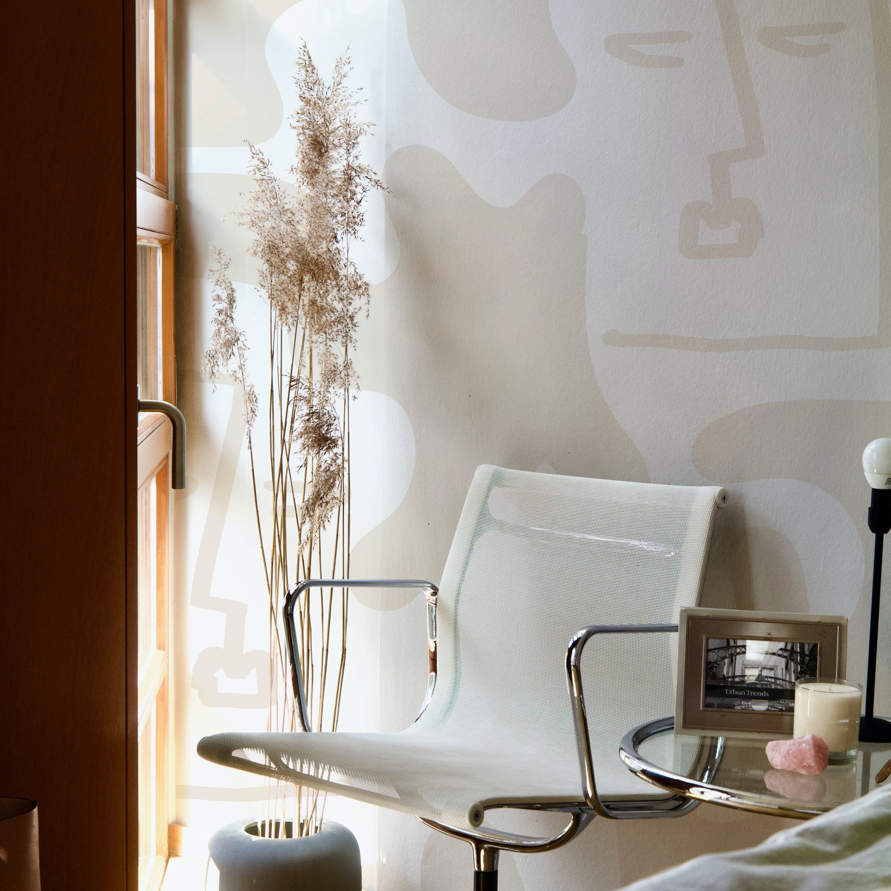 Minimalist Boho Wallpaper featuring abstract linear faces and organic shapes in neutral beige tones, creating a serene and stylish backdrop in a sunlit corner with a modern chair, glass table, and dried plants.