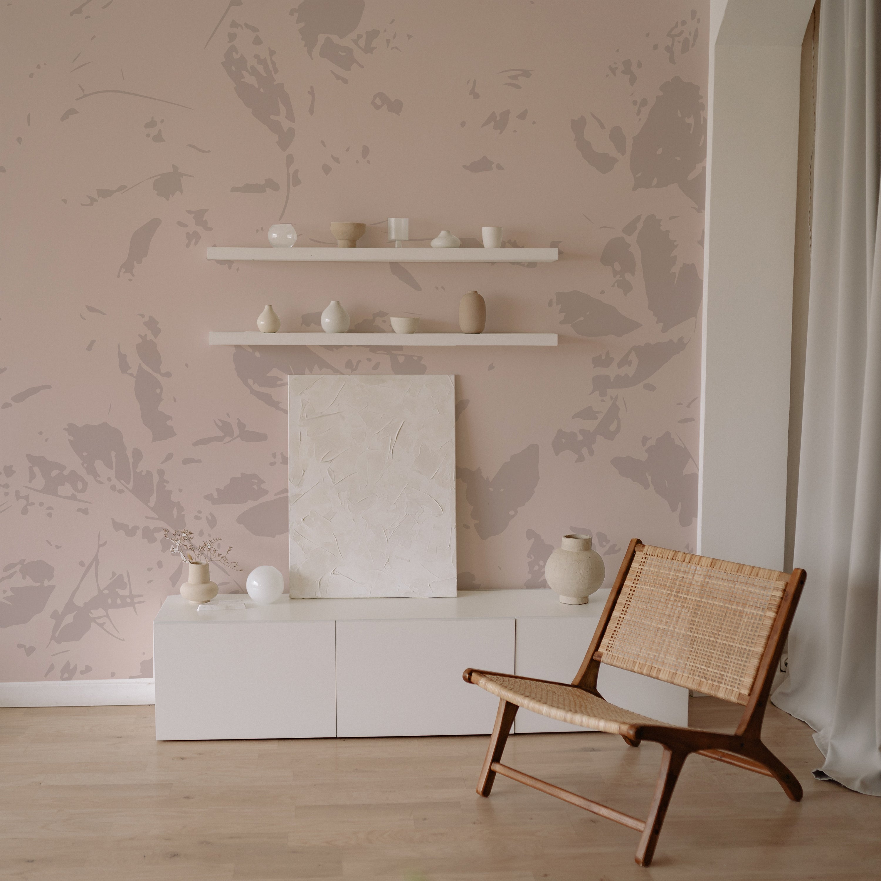 A chic and minimalist room setup showcasing the Abstract Floral - Floral Mural Wallpaper. The wall is adorned with the wallpaper featuring soft gray floral patterns on a blush pink background. The room includes a modern white storage unit, wooden shelves with ceramic decorations, and a stylish wooden armchair, enhancing the elegant and calm feel of the space.
