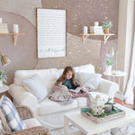 Luxury Watercolor Wall Mural Wallpaper with abstract watercolor shapes in shades of pink and beige with gold accents, adorning the wall of a cozy living room with a white sofa, wicker chair, and stylish decor