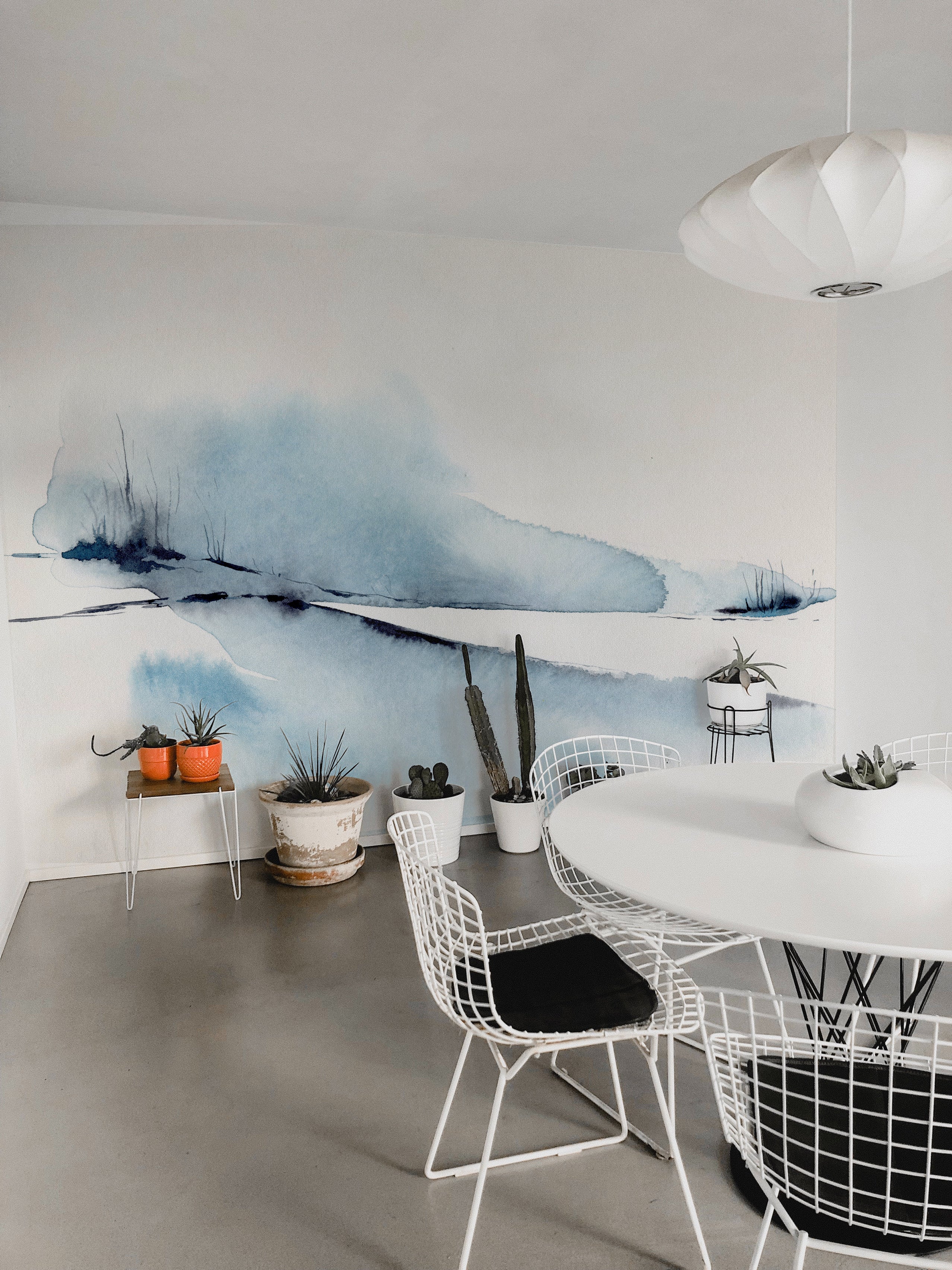 A chic dining area brightened by the "Atmospheric Sky" wall mural. The expansive mural, showcasing vibrant blue hues and abstract watercolor elements, acts as a striking visual centerpiece, contrasting beautifully with the sleek white dining table and wire chairs.