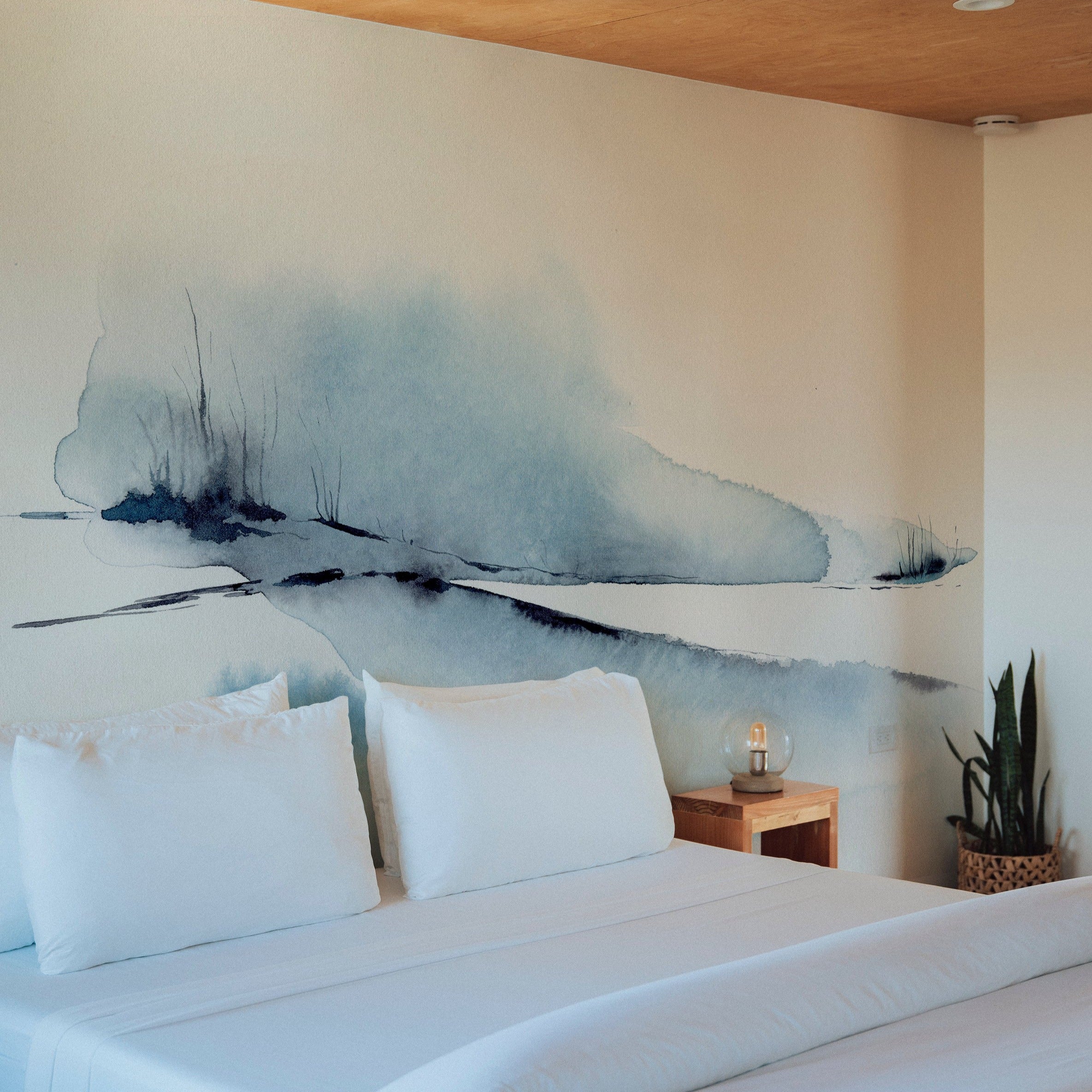 A serene bedroom setup with the "Atmospheric Sky" wall mural painting an ethereal watercolor landscape behind the bed. The mural's soothing blues and detailed brush strokes create a peaceful ambiance, complemented by simple white bedding and minimalistic decor.