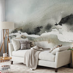 A spacious living room featuring the expansive "Atmospheric Abstraction" wall mural behind a plush beige sofa. The gentle, sweeping brush strokes of the mural in grey and white provide a serene backdrop, complemented by natural wood accents and soft textiles, creating a cozy and inviting environment.