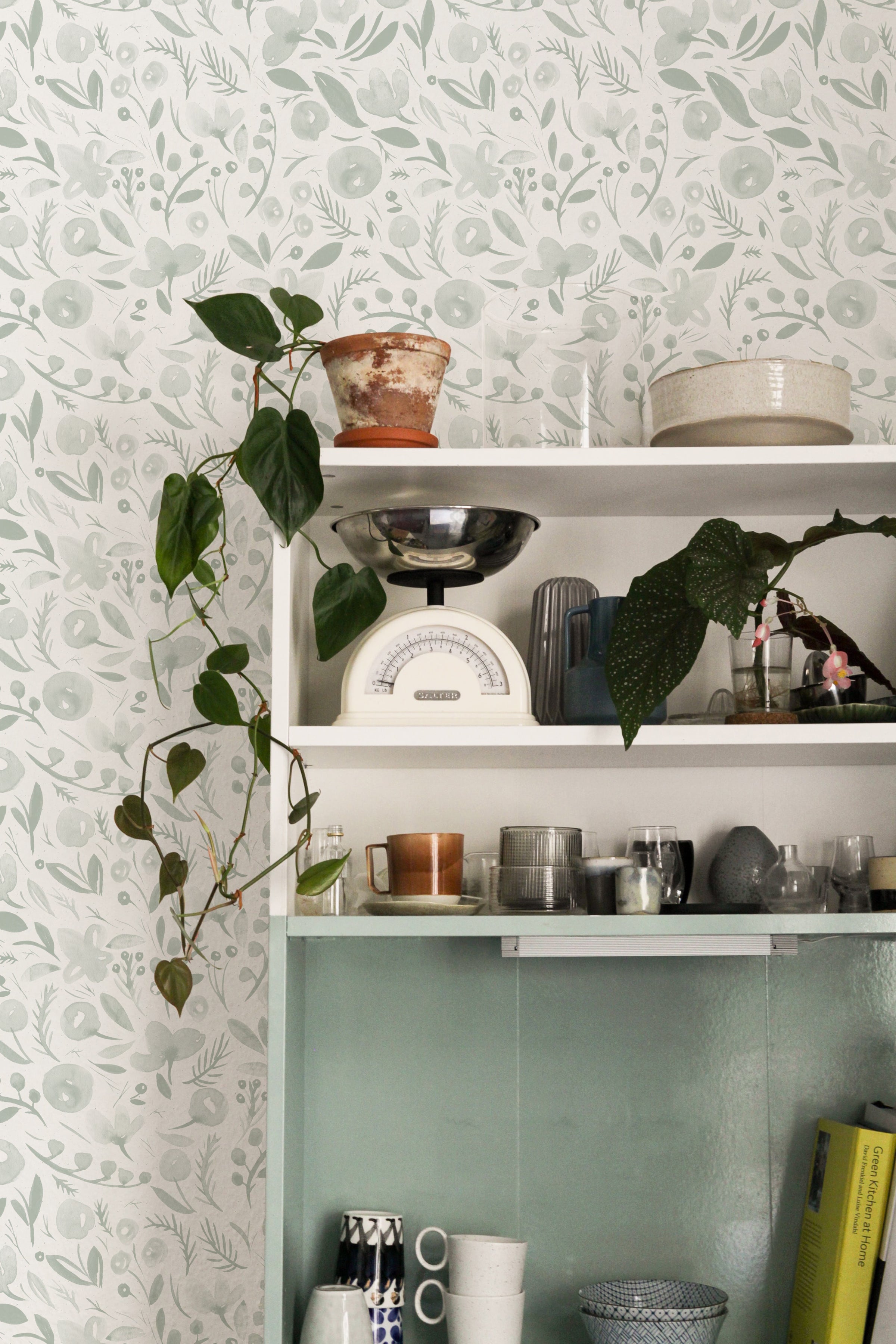 A cozy kitchen corner showcasing the Botanica Wallpaper, adorned with a delicate and elegant botanical pattern in soft shades of green. The wallpaper provides a natural and refreshing backdrop to a white shelving unit, which is decorated with an assortment of household items including a vintage scale, a rustic pot, and various kitchenware, enhancing the serene and homey atmosphere of the space.