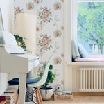 A cozy, well-lit room with a white piano and a green chair. The wall is adorned with Wild Flora Wallpaper, featuring delicate floral arrangements in soft, pastel colors. The window seat is filled with cushions, creating a serene and inviting atmosphere perfect for relaxation.