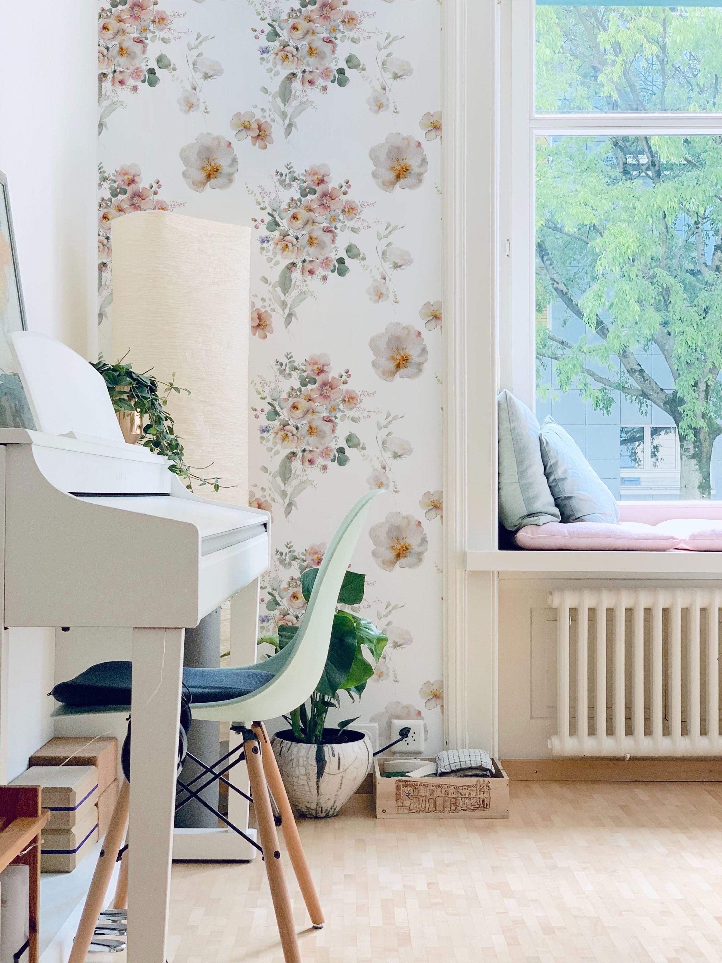 A cozy, well-lit room with a white piano and a green chair. The wall is adorned with Wild Flora Wallpaper, featuring delicate floral arrangements in soft, pastel colors. The window seat is filled with cushions, creating a serene and inviting atmosphere perfect for relaxation.