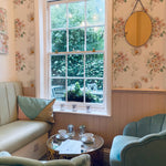 A charming seating area with pastel green chairs and a small round table, complemented by Wild Flora Wallpaper. The floral pattern adds a touch of elegance and tranquility to the room, making it a perfect spot for enjoying a cup of tea and conversation with friends.