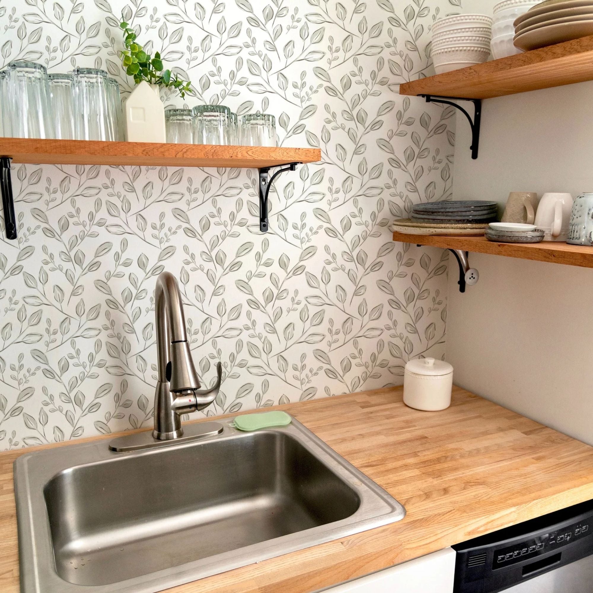 A kitchen corner with a sink under wooden shelves, set against a backdrop of Sweet Watercolour Floral Wallpaper. The gentle floral design on the wallpaper adds a warm and inviting touch to the functional space.