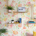 A lively and inviting workspace featuring Bright and Colorful Floral Wallpaper. The wall is decorated with a vibrant watercolor-style floral pattern in shades of pink, yellow, and green. Modern wire wall shelves are mounted against the wallpaper, holding various decorative items and plants, enhancing the room's fresh and cheerful vibe.