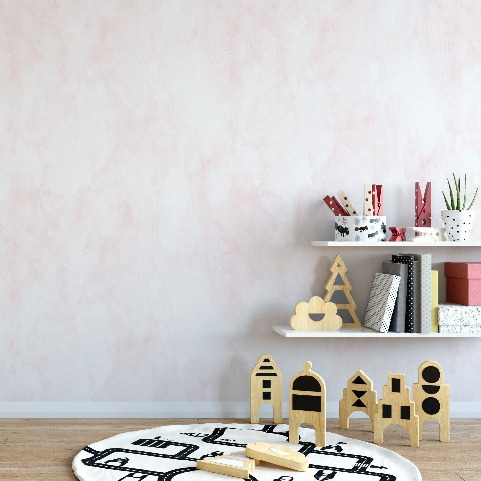 A children's play area with Blush Limewash Wallpaper on the wall. The soft pink texture of the wallpaper creates a serene backdrop, complementing the minimalist shelves with toys, books, and decorative items.